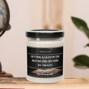 May include: A white candle jar with a black lid and a black label that reads "Smells Like Getting Railed in the Restricted Section by Draco" and "Scented Candle Flare for the Dramatic".