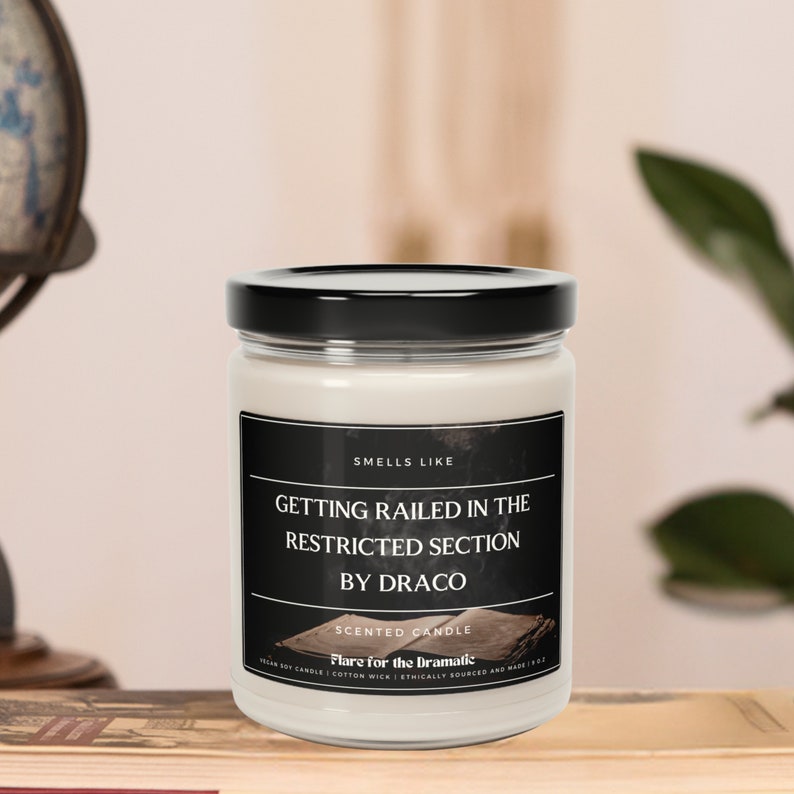 May include: A white candle jar with a black lid and a black label that reads "Smells Like Getting Railed in the Restricted Section by Draco" and "Scented Candle Flare for the Dramatic".