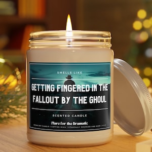 May include: A white candle jar with a black and teal label that reads "Smells Like Getting Fingered in the Fallout by the Ghoul". The label also says "Scented Candle Flare for the Dramatic" and "Vegan Soy Candle | Cotton Wick | Ethically Sourced and Made | 9 oz."