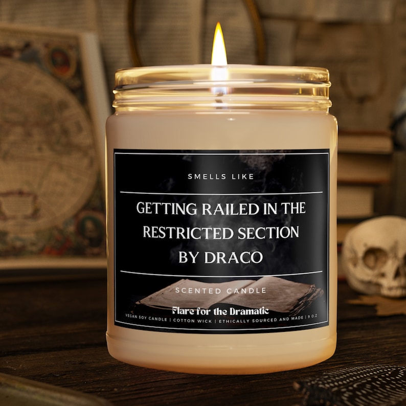 May include: A clear glass candle jar with a black label that reads "SMELLS LIKE GETTING RAILED IN THE RESTRICTED SECTION BY DRACO" and "SCENTED CANDLE Flare for the Dramatic VEGAN SOY CANDLE | COTTON WICK | ETHICALLY SOURCED AND MADE | 9 OZ".