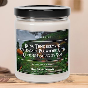May include: A white candle jar with a black lid. The label features a green landscape with a hobbit hole and the text "Smells Like Being Tenderly Fed After-Care Potatoes After Getting Railed By Sam". The text "Scented Candle Flare for the Dramatic" is also on the label.