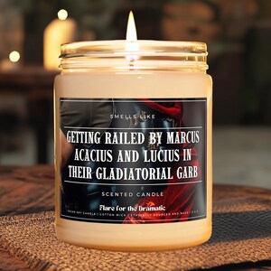 May include: A scented candle with a label that reads "Smells Like Getting Railed by Marcus Acacius and Lucius in Their Gladiatorial Garb". The candle is in a clear glass jar with a gold lid. The label is red, black, and white. The candle is on a brown woven surface.