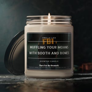May include: A white soy candle with a black label that reads "Smells Like FBI Muffling Your Moans With Booth and Bones" and "Scented Candle Flare for the Dramatic Vegan Soy Candle | Cotton Wick | Ethically Sourced and Made in the US"