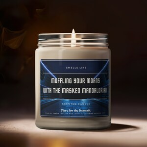 May include: A white soy candle in a glass jar with a blue and black label that reads "Smells Like Muffling Your Moans With The Masked Mandalorian" and "Scented Candle Flare for the Dramatic".