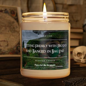 Op de afbeelding: Een bruine glazen kaars pot met een wit label. Het label heeft een groene en bruine afbeelding van een hobbitgat met de tekst "Smells Like Getting Freaky With Frodo And Banged In Bag End". Het label vermeldt ook "Scented Candle Flare for the Dramatic Soy Candle | Cotton Wick | Ethically Sourced and Made | 9 oz"
