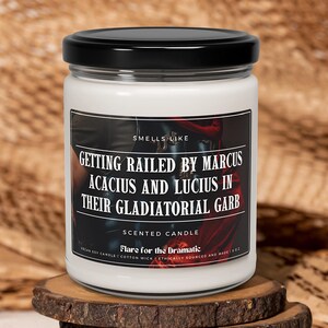 May include: A white candle with a black lid. The label reads "Smells Like Getting Railed by Marcus Acacius and Lucius in Their Gladiatorial Garb". The candle is a scented candle and says "Flare for the Dramatic".