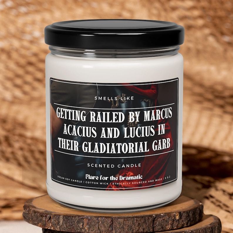 May include: A white candle with a black lid. The label reads "Smells Like Getting Railed by Marcus Acacius and Lucius in Their Gladiatorial Garb". The candle is a scented candle and says "Flare for the Dramatic".