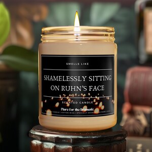 May include: A brown glass candle with a black label that reads "Smells Like Shamelessly Sitting On Ruhn's Face" and "Scented Candle Flare for the Dramatic".
