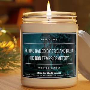 Puede incluir: Una vela de soja blanca con una etiqueta negra que dice "Smells Like Getting Railed by Eric and Bill in the Bon Temps Cemetery." La etiqueta también incluye el texto "Scented Candle" y "Flare for the Dramatic."