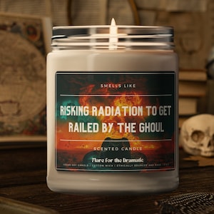 May include: A beige candle jar with a black label that reads "Smells Like Risking Radiation to Get Railed by the Ghoul". The label also includes the text "Scented Candle Flare for the Dramatic Vegan Soy Candle | Cotton Wick | Ethically Sourced and Made | 8 oz."