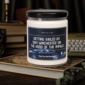 May include: A white candle with a black lid and a label that reads "Smells Like Getting Railed By Sam Winchester On The Hood Of The Impala" with a blue and black image of a car. The label also says "Scented Candle" and "Flare for the Dramatic".