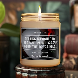 Könnte beinhalten: Eine braune Glaskerze mit einem weißen Etikett, auf dem "Smells Like Getting Ravished by the Phantom in His Crypt Under the Opera House" steht. Die Kerze ist angezündet und hat eine rote Rose auf dem Etikett.