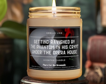 May include: A brown glass candle with a white label that reads "Smells Like Getting Ravished by the Phantom in His Crypt Under the Opera House". The candle is lit and has a red rose on the label.