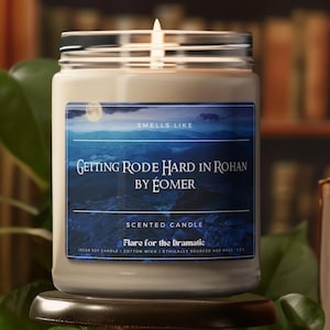 Peut inclure: Une bougie parfumée allumée dans un bocal en verre avec un couvercle argenté. L'étiquette indique "Getting Rode Hard in Rohan by Éomer" avec une scène de montagne. La bougie est étiquetée "Flare for the Dramatic".