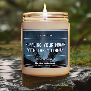 Può includere: Un barattolo di vetro marrone con una candela e un'etichetta nera su cui è scritto "SMELLS LIKE MUFFLING YOUR MOANS WITH THE MOTHMAN" e "SCENTED CANDLE Flare for the Dramatic" su sfondo bianco. La candela è accesa e la fiamma è visibile.