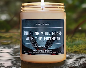 May include: A brown glass candle jar with a black label that reads "SMELLS LIKE MUFFLING YOUR MOANS WITH THE MOTHMAN" and "SCENTED CANDLE Flare for the Dramatic" with a white background. The candle is lit and the flame is visible.
