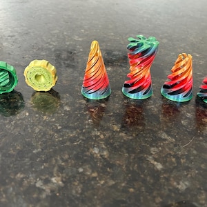 May include: A set of six colourful, 3D printed, geometric shapes. The shapes are a combination of green, yellow, red, and black. The shapes are arranged in a row on a dark grey surface.