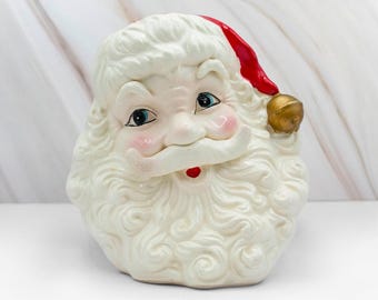 May include: A ceramic Santa Claus head with a red hat and a gold bell. The Santa has a white beard and mustache, rosy cheeks, and blue eyes. The decorative piece is a festive holiday item.