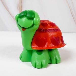 May include: A green turtle figurine with a red shell decorated with heart shapes. The turtle has closed eyes and a smiling mouth. The figurine is made of plastic and is on a white surface.