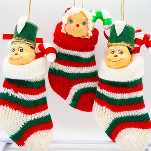 May include: Three Christmas stocking ornaments with elf and Santa Claus faces. The stockings are red, green, and white striped, with white cuffs. Each ornament has a candy cane and a gold string for hanging.