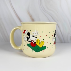 May include: Cream-colored ceramic mug featuring Mickey Mouse in a festive Christmas design. Mickey is depicted on a green sled, wearing a Santa hat, with red and green polka dots. The mug has a rounded shape and a large handle.