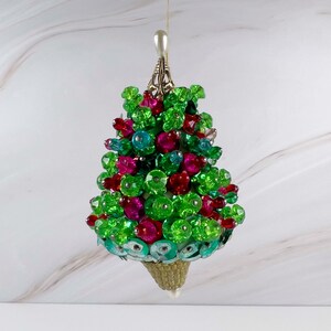 May include: A cone-shaped hanging ornament covered in vibrant green, fuchsia, and turquoise faceted beads and sequins. The ornament features a metallic silver-toned cap at the top and a textured, tan-colored conical base at the bottom.