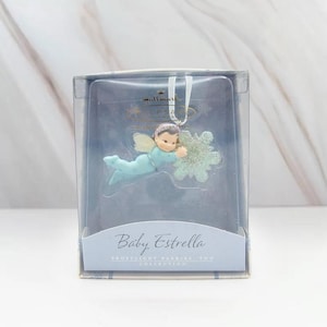 May include: A Hallmark Baby Estrella Christmas ornament. The ornament features a baby angel in a light blue outfit with a snowflake. The ornament is in a clear plastic box with the words "Baby Estrella" on the front.