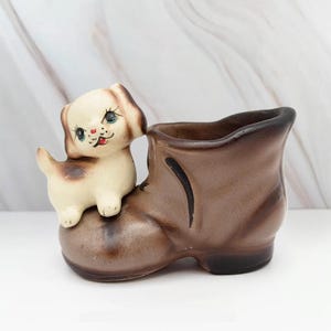 May include: A vintage ceramic planter shaped like a brown boot with a small, cream-colored puppy figurine perched on top. The puppy has brown spots and a red nose. The boot has a dark brown interior.