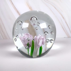 May include: A clear glass paperweight with two lavender-colored tulip flowers and green stems encased inside. The glass sphere has several air bubbles throughout. The paperweight is on a white surface with a marble-patterned background.