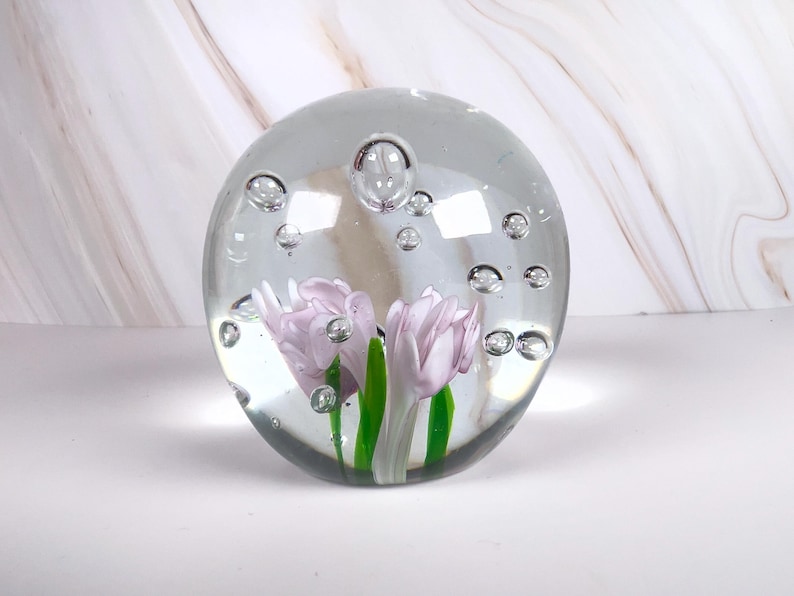 May include: A clear glass paperweight with two lavender-colored tulip flowers and green stems encased inside. The glass sphere has several air bubbles throughout. The paperweight is on a white surface with a marble-patterned background.