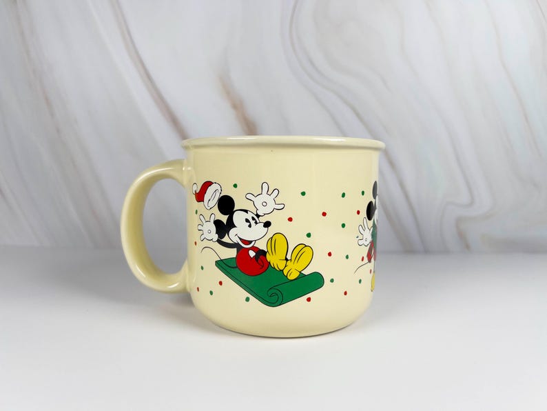 May include: Cream-colored ceramic mug featuring Mickey Mouse in a festive Christmas design. Mickey is depicted on a green sled, wearing a Santa hat, with red and green polka dots. The mug has a rounded shape and a large handle.