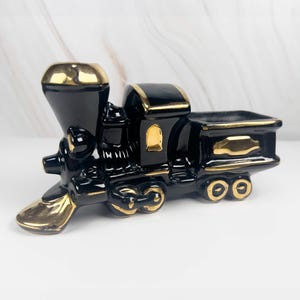 May include: Black and gold ceramic train figurine. The train features a detailed design with gold accents on the smokestack, front, and wheels. The train is a decorative item, likely for display or as a collectible.