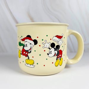 May include: Cream-colored ceramic mug featuring two cartoon mice in festive attire. One mouse is ice skating, and the other is waving. Both mice wear Santa hats and red, green, and yellow accents. The mug has a large handle.