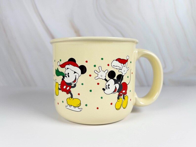 May include: Cream-colored ceramic mug featuring two cartoon mice in festive attire. One mouse is ice skating, and the other is waving. Both mice wear Santa hats and red, green, and yellow accents. The mug has a large handle.