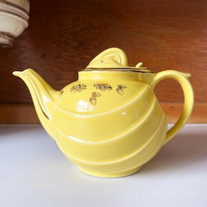 May include: A yellow ceramic teapot with a curved spout and handle. The teapot has a lid with a swirl design and is decorated with gold leaf patterns. The teapot has a ribbed design and is sitting on a white surface.