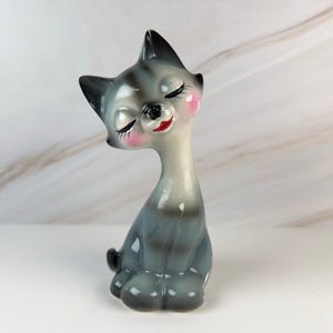 May include: A ceramic cat figurine with a glossy finish, featuring a grey and white gradient body, pointed ears, and a long neck. The cat has painted closed eyes with long eyelashes, bright pink cheeks, and red lips, sitting in an upright position.