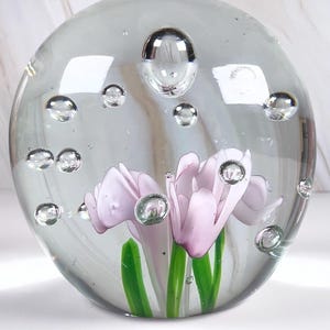 May include: A clear glass paperweight with a spherical shape. Inside, delicate pink flowers with green stems are encased, surrounded by air bubbles. The paperweight is a decorative item.