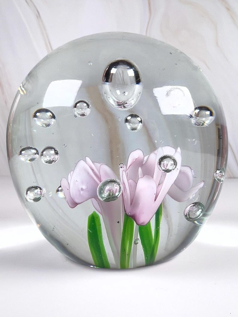 May include: A clear glass paperweight with a spherical shape. Inside, delicate pink flowers with green stems are encased, surrounded by air bubbles. The paperweight is a decorative item.