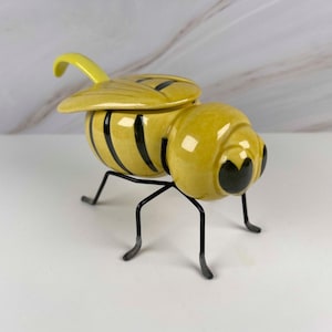 May include: A ceramic bee-shaped container with a yellow and black striped body and a removable lid. The insect-themed vessel features a glossy finish, dark eyes, and is supported by four thin, black metal legs that elevate the main body.