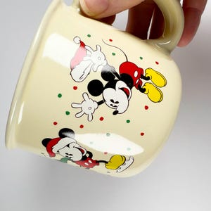 May include: Cream-colored ceramic mug featuring Mickey Mouse in a festive Christmas design. Mickey is depicted in a Santa hat and red shorts, with colorful polka dots. The mug has a curved shape and a matching handle.