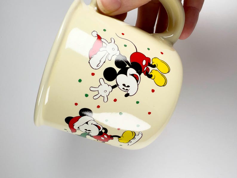 May include: Cream-colored ceramic mug featuring Mickey Mouse in a festive Christmas design. Mickey is depicted in a Santa hat and red shorts, with colorful polka dots. The mug has a curved shape and a matching handle.