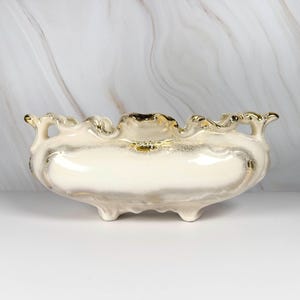 May include: Cream-colored ceramic decorative bowl with gold trim. The oval-shaped bowl has a scalloped rim and sits on three small feet. The interior and trim feature gold accents, creating an elegant, vintage aesthetic.