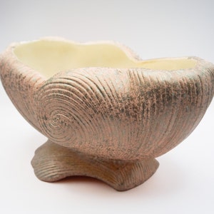 May include: A large, ceramic planter shaped like a seashell with a light pink and green glaze. The planter has a wide, shallow bowl and a rounded base.