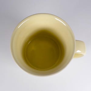 May include: Overhead view of a pale yellow ceramic mug with a matching handle. The interior of the mug is a darker shade of yellow. The mug is set against a white background.