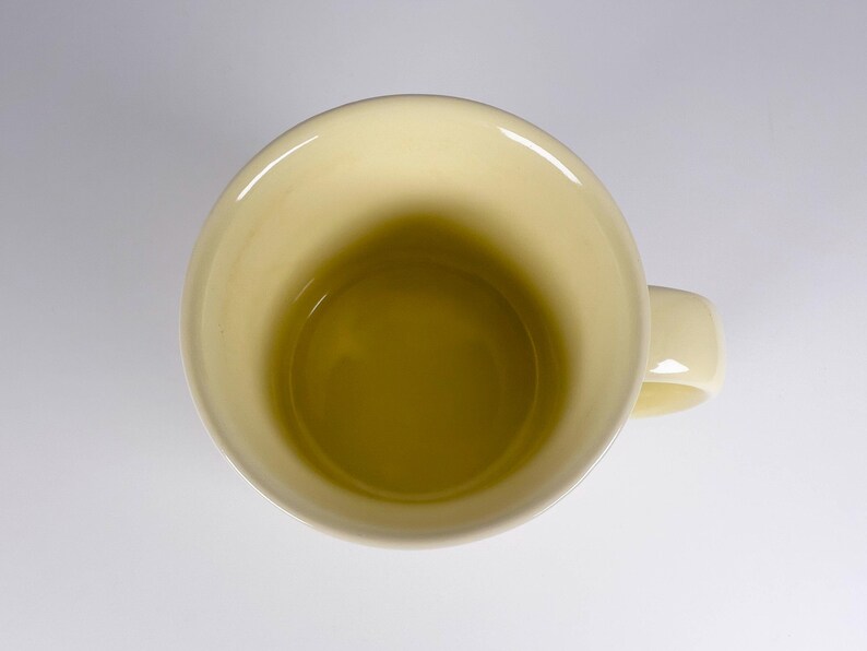 May include: Overhead view of a pale yellow ceramic mug with a matching handle. The interior of the mug is a darker shade of yellow. The mug is set against a white background.