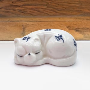 May include: A white ceramic cat figurine in a curled-up sleeping position. The cat has closed eyes and is decorated with small blue floral accents. The figurine is on a white surface with a wooden background.