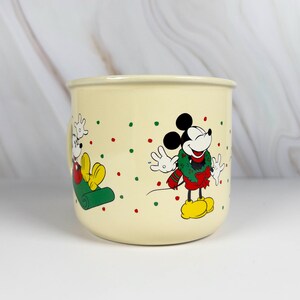 May include: Cream-colored ceramic mug featuring Mickey Mouse in a Christmas design. Mickey is depicted in a red and green outfit with a green wreath. The mug is decorated with red and green polka dots.