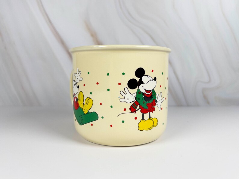 May include: Cream-colored ceramic mug featuring Mickey Mouse in a Christmas design. Mickey is depicted in a red and green outfit with a green wreath. The mug is decorated with red and green polka dots.