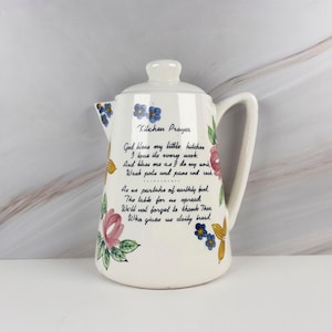 May include: A cream-colored ceramic pitcher with a lid and handle, featuring hand-painted floral motifs in pink, blue, and yellow. The front displays the text Kitchen Prayer followed by a poem about blessing the kitchen, cooking, and daily bread.