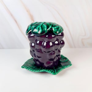 May include: A glossy ceramic container shaped like a cluster of dark purple grapes. The lid features a sculpted green leaf design, and the base is a matching green leaf-shaped saucer. The surface has a reflective, smooth, and glazed finish.